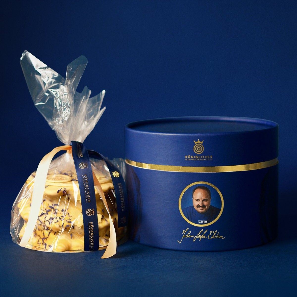 Baumkuchen der Johann Lafer Edition in Klarsichtfolie neben der blauen Geschenkdose